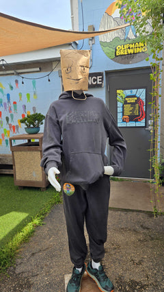 Person wearing a gray hoodie and pants with a paper bag over their head, standing in front of a building with a sign that reads 'Oliphant Brewing'.